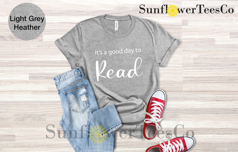 Op de afbeelding: Lichtgrijs heather t-shirt met de tekst "It's a good day to Read" in witte letters. Het shirt wordt gecombineerd met lichtblauwe jeans en rode canvas sneakers met witte veters. De afbeelding is een flat lay.