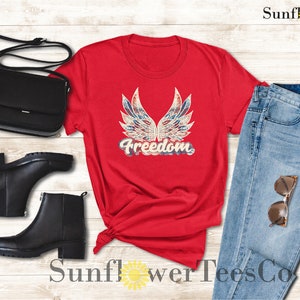 Puede incluir: Camiseta roja con un dise&ntilde;o de bandera estadounidense desgastado y la palabra "Freedom" en una fuente vintage.