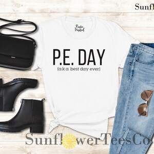 Op de afbeelding: Wit T-shirt met zwarte tekst die "P.E. DAY (a.k.a. best day ever)" luidt. Het shirt is gestyled met een blauwe jeans, zwarte laarzen en een zwarte handtas.