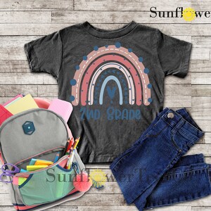 May include: A dark grey t-shirt with a rainbow design and the text "2nd Grade". The rainbow is made up of pink, red, blue, and white stripes with white polka dots and blue stars. The shirt is laid out on a wooden surface with a backpack, jeans, and school supplies.
