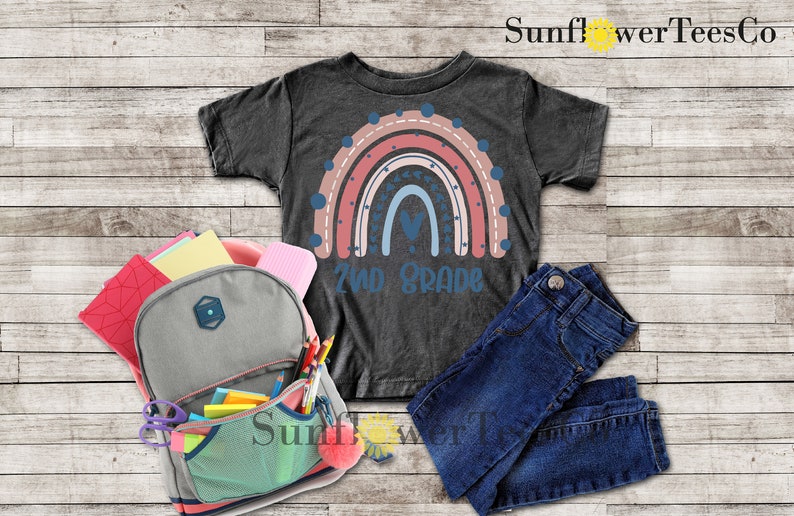 May include: A dark grey t-shirt with a rainbow design and the text "2nd Grade". The rainbow is made up of pink, red, blue, and white stripes with white polka dots and blue stars. The shirt is laid out on a wooden surface with a backpack, jeans, and school supplies.