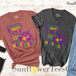 May include: Two t-shirts with a Mardi Gras theme. The left shirt is a rust color with a graphic of the state of Louisiana with the words "Louisiana", "NOLA", and "Mardi Gras". The right shirt is a dark gray with a similar graphic but with the words "Louisiana" and "Mardi Gras".