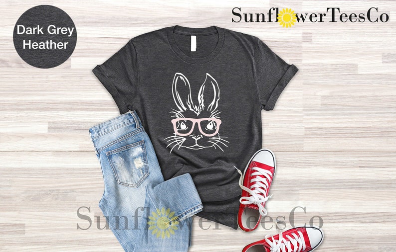 May include: Dark grey heather t-shirt with a white graphic of a bunny wearing pink glasses. The t-shirt is laid flat on a wooden surface with a pair of blue jeans and red sneakers.