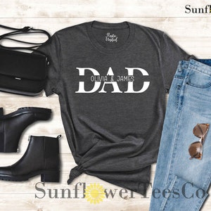 Op de afbeelding: Een donkergrijs T-shirt met het woord "DAD" in witte blokletters. De tekst "Olivia & James" is in kleinere witte letters onder het woord "DAD" gedrukt.