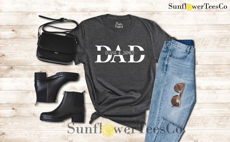 Op de afbeelding: Een donkergrijs T-shirt met het woord "DAD" in witte blokletters. De tekst "Olivia & James" is in kleinere witte letters onder het woord "DAD" gedrukt.