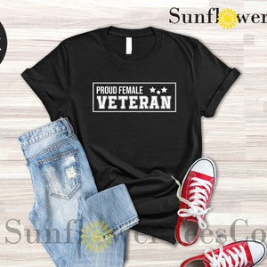 May include: Black t-shirt with white text that reads "Proud Female Veteran" with three stars above the text. The shirt is laid flat on a wood surface with a pair of blue jeans and red sneakers.
