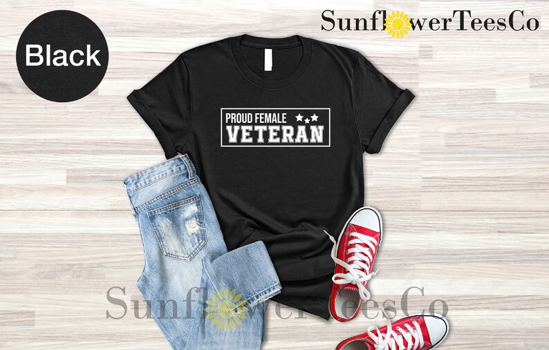 May include: Black t-shirt with white text that reads "Proud Female Veteran" with three stars above the text. The shirt is laid flat on a wood surface with a pair of blue jeans and red sneakers.