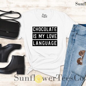 May include: White t-shirt with the text "CHOCOLATE IS MY LOVE LANGUAGE" in a black rectangular box. The image also includes black ankle boots, a black purse, and blue jeans with sunglasses.
