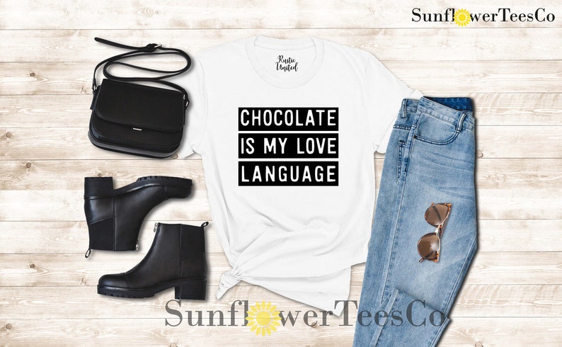 May include: White t-shirt with the text "CHOCOLATE IS MY LOVE LANGUAGE" in a black rectangular box. The image also includes black ankle boots, a black purse, and blue jeans with sunglasses.