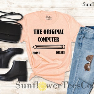 K&ouml;nnte beinhalten: Ein pfirsichfarbenes T-Shirt mit dem Text "THE ORIGINAL COMPUTER" und einem Bleistift mit den W&ouml;rtern "PRINT" und "DELETE" darunter. Das Shirt ist mit einer blauen Jeans, schwarzen Stiefeln und einer schwarzen Handtasche gestylt.