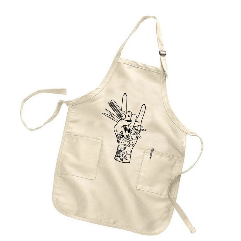 May include: A cream-colored apron with two front pockets and a pen slot. The apron features a black graphic of a hand making a rock-and-roll gesture, holding scissors, a comb, and a tattoo design.