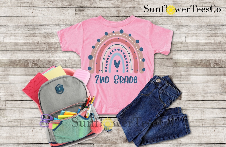 May include: A pink t-shirt with a rainbow design and the text "2nd Grade". The rainbow is made up of different shades of pink, orange, and blue. There are small hearts and dots around the rainbow. The shirt is laid out on a wooden surface with a backpack, pencils, and jeans.