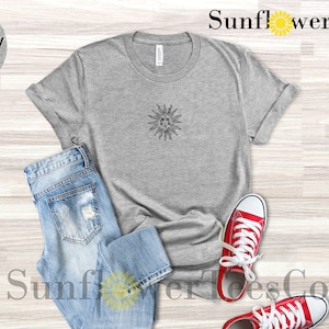 May include: A light grey heather t-shirt with a black sun graphic. The shirt is laid flat on a wooden surface with a pair of blue jeans and red sneakers.