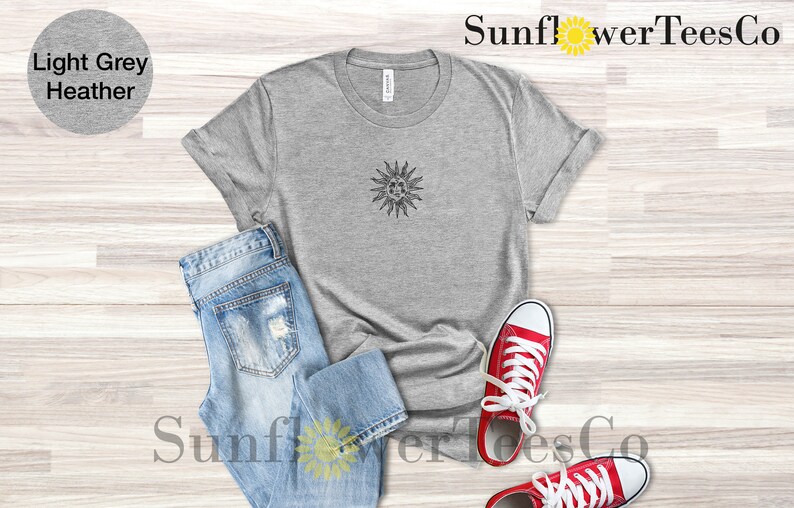 May include: A light grey heather t-shirt with a black sun graphic. The shirt is laid flat on a wooden surface with a pair of blue jeans and red sneakers.