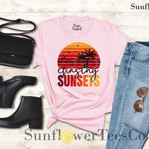 May include: Pink t-shirt with a retro sunset graphic and the text "chasing sunsets". The shirt is paired with blue jeans, black boots, and a black purse.