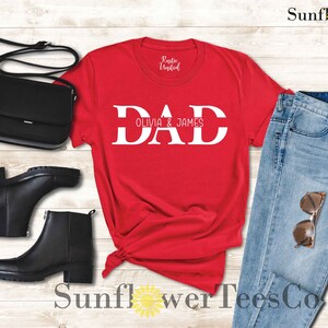 Op de afbeelding: Rood T-shirt met witte tekst die "DAD Olivia & James" zegt. Het shirt ligt op een houten oppervlak met een paar zwarte laarzen, een zwarte tas en een blauwe jeans.