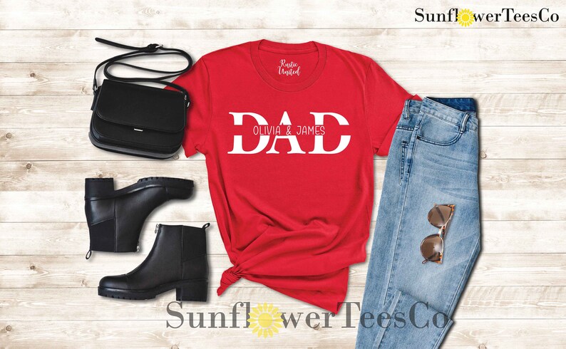 Op de afbeelding: Rood T-shirt met witte tekst die "DAD Olivia & James" zegt. Het shirt ligt op een houten oppervlak met een paar zwarte laarzen, een zwarte tas en een blauwe jeans.