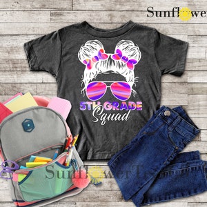 May include: A dark grey t-shirt with a white graphic of a girl with pigtails wearing sunglasses and the text "5th Grade Squad".