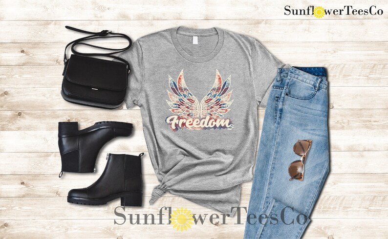 Puede incluir: Una camiseta gris con un gr&aacute;fico desgastado de alas rojas, blancas y azules con la palabra "Freedom" en una fuente de estilo acuarela descolorida.