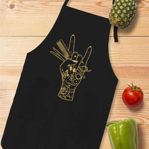 May include: Black apron with a gold-colored hand graphic holding scissors, a comb, and other barber tools. The hand is in a rock-and-roll gesture. A pineapple, tomato, and green bell pepper are on a wooden surface.