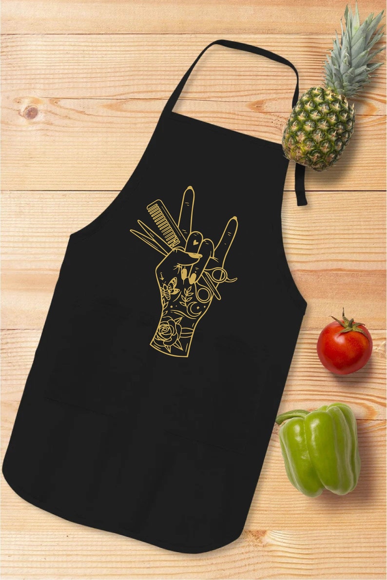 May include: Black apron with a gold-colored hand graphic holding scissors, a comb, and other barber tools. The hand is in a rock-and-roll gesture. A pineapple, tomato, and green bell pepper are on a wooden surface.