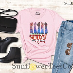 May include: Pink t-shirt with a graphic of raised fists in front of a map of the United States. The graphic is colorful and includes the text "Hispanic Heritage Month".