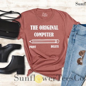 K&ouml;nnte beinhalten: Ein rostiges T-Shirt mit dem Text "The Original Computer" und einer Bleistiftgrafik mit den W&ouml;rtern "Print" und "Delete" darunter. Das Shirt wird mit einer blauen Jeans, einer schwarzen Handtasche und schwarzen Stiefeletten kombiniert.