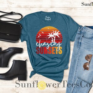 May include: A teal blue t-shirt with a vintage sunset graphic and the text "chasing sunsets". The shirt is paired with blue jeans, black ankle boots, and a black crossbody bag.