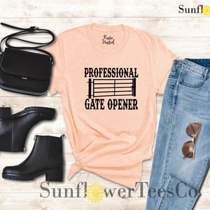 Op de afbeelding: Een perzikkleurig T-shirt met de tekst "Professional Gate Opener" en een zwart silhouet van een poort. Het shirt is gestyled met een blauwe jeans, zwarte laarzen en een zwarte tas.