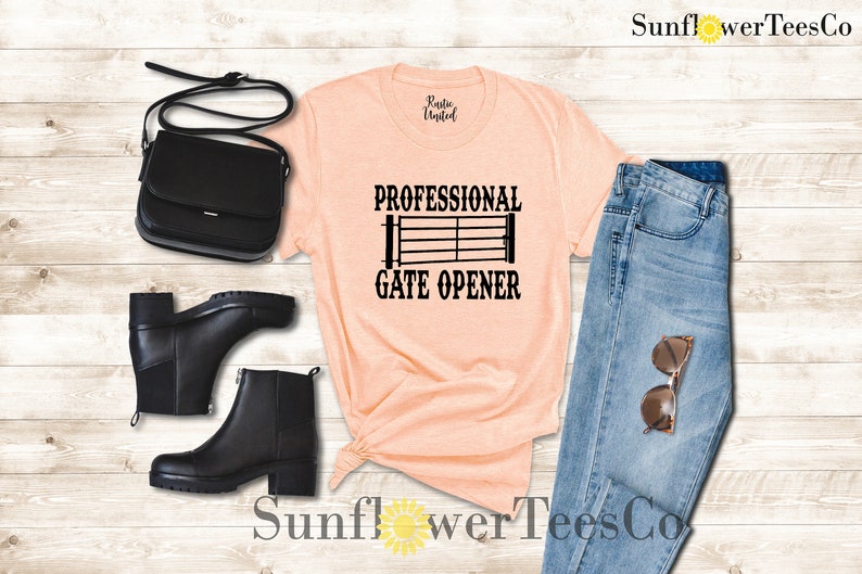 Op de afbeelding: Een perzikkleurig T-shirt met de tekst "Professional Gate Opener" en een zwart silhouet van een poort. Het shirt is gestyled met een blauwe jeans, zwarte laarzen en een zwarte tas.