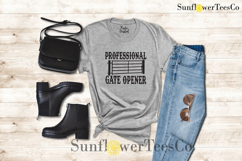 Op de afbeelding: Een grijs T-shirt met de tekst "Professional Gate Opener" en een zwart silhouet van een poort. Het shirt ligt op een houten oppervlak met een paar zwarte laarzen, een zwarte tas en een blauwe jeans.