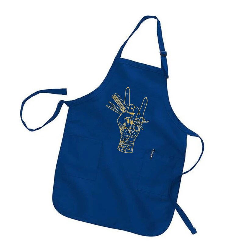 May include: A royal blue apron with two front pockets and a neck strap. A gold-colored graphic of a hand with scissors, a comb, and other barber tools is printed on the front. The apron is designed for use in a salon or barber shop.