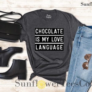 May include: Dark gray t-shirt with the text "CHOCOLATE IS MY LOVE LANGUAGE" in a black and white design. The shirt is styled with black ankle boots, a black purse, and light blue jeans with sunglasses.