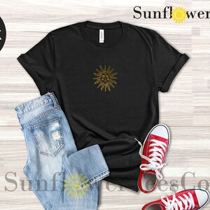 May include: Black t-shirt with a gold sun graphic. The t-shirt is laid flat on a wooden surface with a pair of blue jeans and red sneakers.