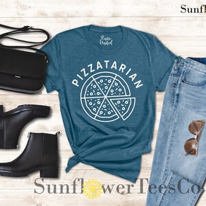 May include: A teal blue t-shirt with the text "PIZZATARIAN" in white lettering and a white outline of a pizza with slices. The shirt is laying on a wooden surface with a pair of black boots, a black purse, and blue jeans.