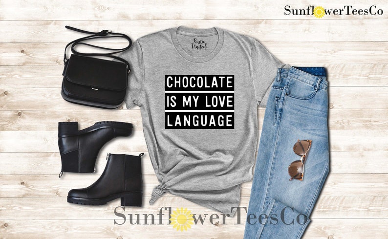 May include: A gray t-shirt with the text "CHOCOLATE IS MY LOVE LANGUAGE" in a black and white design. The image also includes a black purse, black boots, and blue jeans with sunglasses.