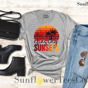 May include: A gray t-shirt with a retro sunset graphic and the text "chasing sunsets". The shirt is paired with blue jeans, black boots, and a black purse.