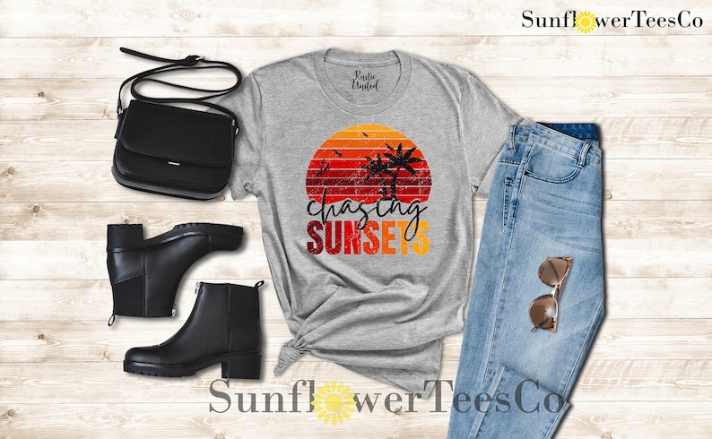 May include: A gray t-shirt with a retro sunset graphic and the text "chasing sunsets". The shirt is paired with blue jeans, black boots, and a black purse.