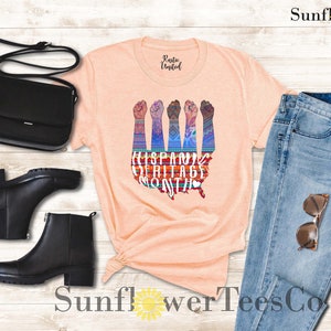 May include: Peach colored t-shirt with a graphic of raised fists in front of a stylized map of the United States. The graphic is made up of colorful patterns and the text "Hispanic Heritage Month".