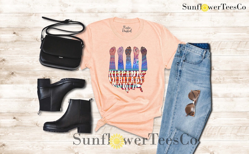 May include: Peach colored t-shirt with a graphic of raised fists in front of a stylized map of the United States. The graphic is made up of colorful patterns and the text "Hispanic Heritage Month".