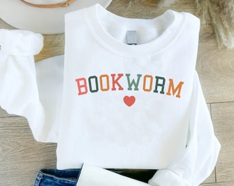 Puede incluir: Sudadera blanca con el texto "BOOKWORM" en letras mayúsculas de colores y un pequeño corazón rojo.