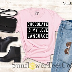 May include: A light pink t-shirt with the text "CHOCOLATE IS MY LOVE LANGUAGE" in black. The shirt is styled with black ankle boots, a black purse, and blue jeans with sunglasses.