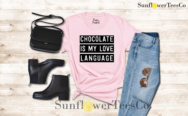 May include: A light pink t-shirt with the text "CHOCOLATE IS MY LOVE LANGUAGE" in black. The shirt is styled with black ankle boots, a black purse, and blue jeans with sunglasses.