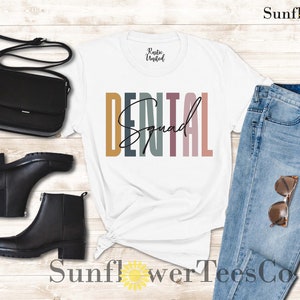 May include: White t-shirt with the text "DENTAL Squad" in colorful vertical stripes. The shirt is styled with a black crossbody bag, black ankle boots, and blue jeans.