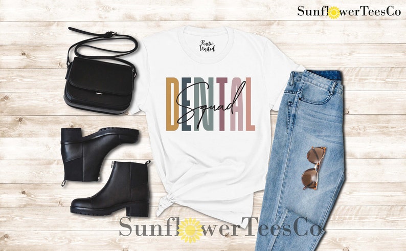 May include: White t-shirt with the text "DENTAL Squad" in colorful vertical stripes. The shirt is styled with a black crossbody bag, black ankle boots, and blue jeans.