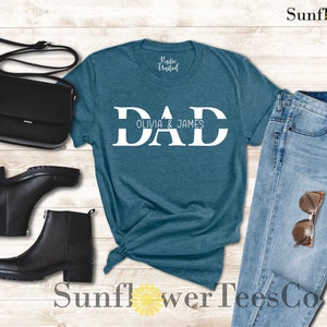 Op de afbeelding: Een tealblauw t-shirt met het woord "DAD" in witte blokletters en de tekst "Olivia & James" eronder. Het shirt wordt gecombineerd met een blauwe jeans, zwarte laarzen en een zwarte tas.