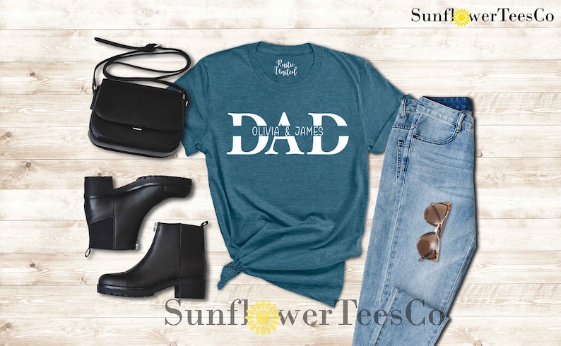 Op de afbeelding: Een tealblauw t-shirt met het woord "DAD" in witte blokletters en de tekst "Olivia & James" eronder. Het shirt wordt gecombineerd met een blauwe jeans, zwarte laarzen en een zwarte tas.