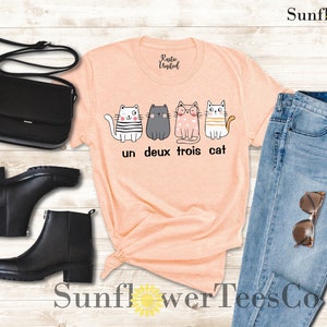 以下が含まれることがあります： 桃色のTシャツに、4匹の猫のグラフィックと「un deux trois cat」の文字がプリントされています。Tシャツは、青いジーンズ、黒いブーツ、黒いショルダーバッグと合わせています。