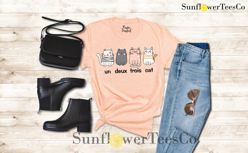 以下が含まれることがあります： 桃色のTシャツに、4匹の猫のグラフィックと「un deux trois cat」の文字がプリントされています。Tシャツは、青いジーンズ、黒いブーツ、黒いショルダーバッグと合わせています。