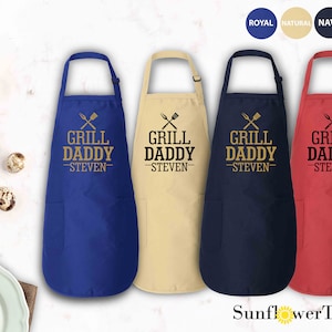 May include: Four aprons in different colors: royal blue, natural beige, navy blue, and red. Each apron has a pocket and the text "Grill Daddy -Steven-  " with crossed grilling utensils.